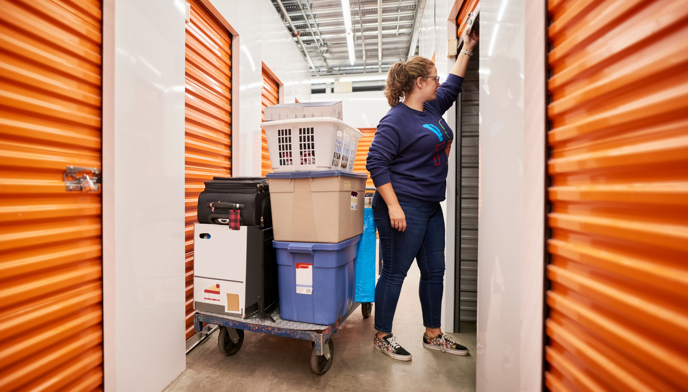long term storage solutions - Woman opening the shutter of a self storage unit with a trolley full of goods and items in the aisle of self-storage building