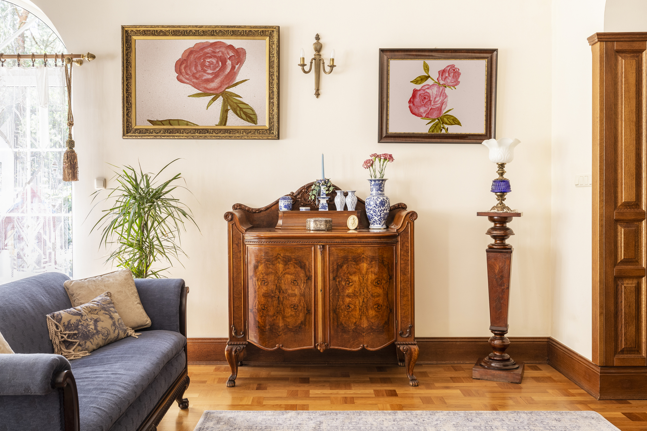 antique cabinet with porcelain decorations, paintings with roses and blue sofa in a living room interior