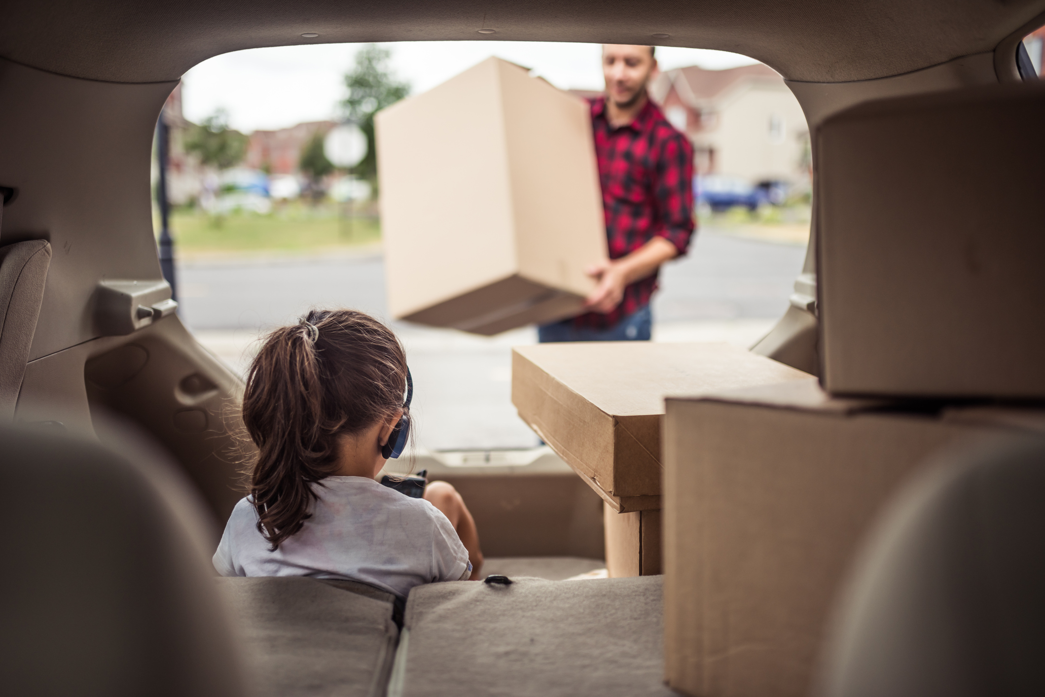 Little girl playing and listening to music in the trunk of the car while her father is loading moving boxes into the car