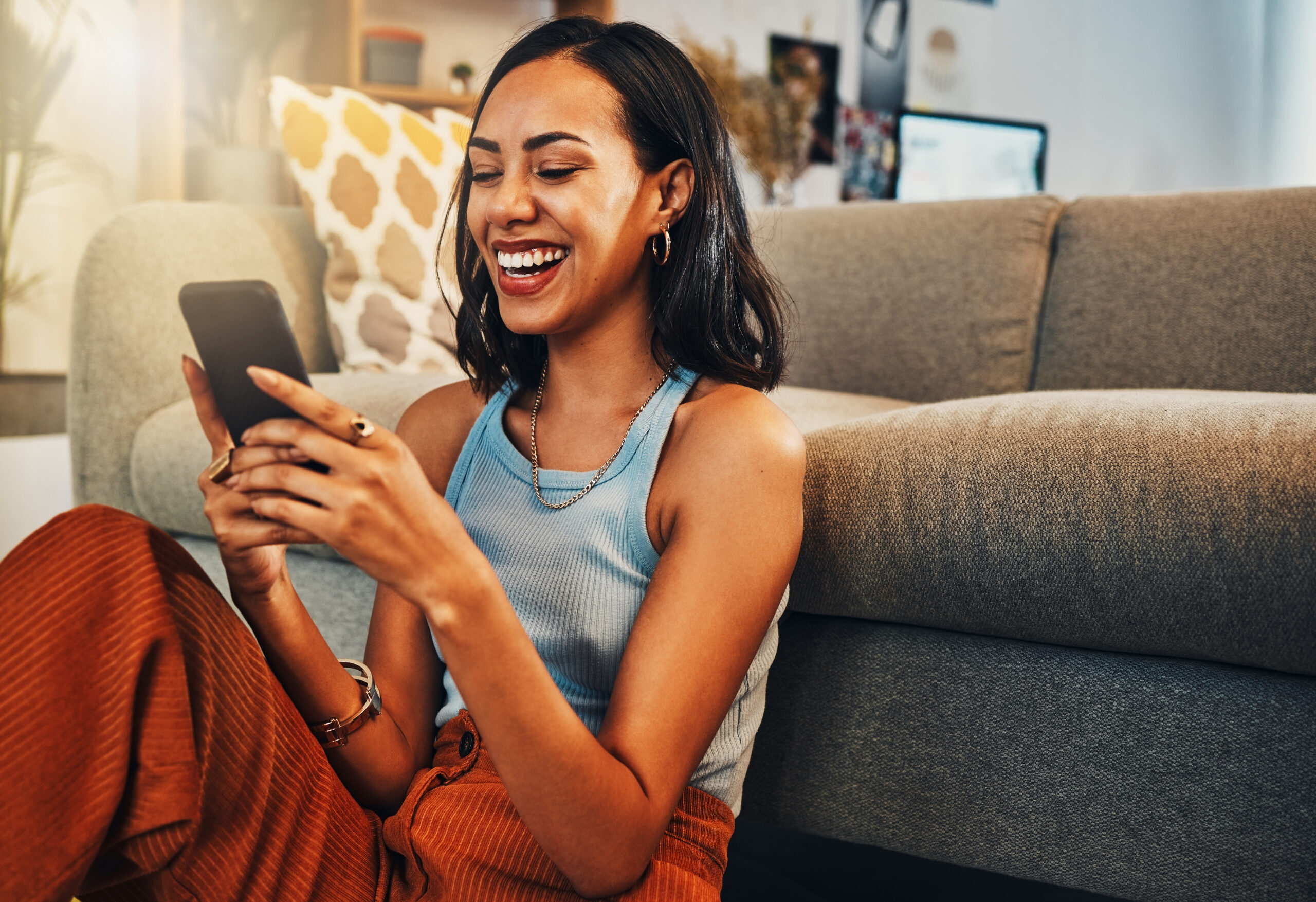 Young woman sitting on the floor in front of couch smiling and looking at mobile phone