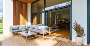 Wooden patio connected to a private building, with seating area and cozy lounge zone, open window with entrance to the house.